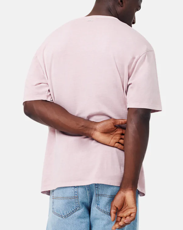 A person in an oversized light pink Industrie The Del Sur Tee and blue jeans stands with their back to the camera, one hand holding the other behind their back.