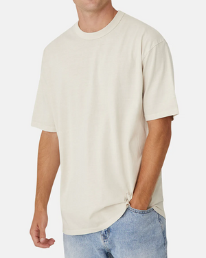 A person wearing the oversized Industrie The Del Sur Tee in off-white and light blue jeans with one hand in their pocket stands against a white background, highlighting the casual 100% cotton outfit. The person's head is not shown in the image.