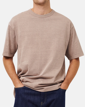 A person wearing an oversized beige Industrie The Del Sur Tee and dark blue acid wash jeans stands with hands in pockets against a plain white background.