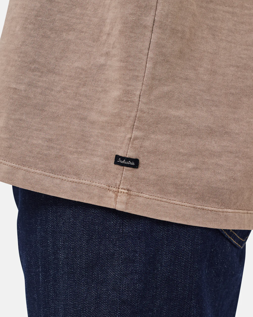 Close-up of the bottom hem of the Industrie The Del Sur Tee, a beige 100% cotton oversized t-shirt with a small black "Industrie" label, styled over dark blue denim jeans.