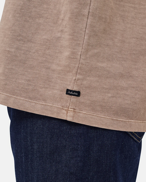 Close-up of the bottom hem of the Industrie The Del Sur Tee, a beige 100% cotton oversized t-shirt with a small black "Industrie" label, styled over dark blue denim jeans.