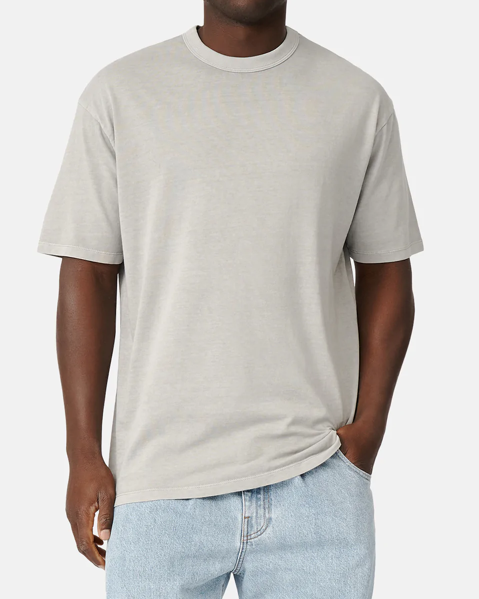 A person in the Industrie The Del Sur Tee, a light gray, 100% cotton casual oversized fit shirt, paired with light blue jeans, stands against a white background. With one hand pocketed and the other hanging casually by their side.