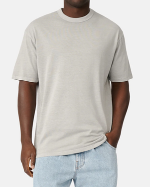 A person in the Industrie The Del Sur Tee, a light gray, 100% cotton casual oversized fit shirt, paired with light blue jeans, stands against a white background. With one hand pocketed and the other hanging casually by their side.