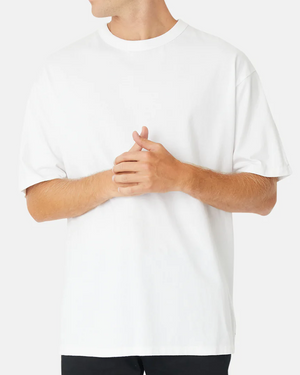 A person wearing an oversized Industrie The Del Sur Tee in white and black pants stands against a light gray background, with hands clasped in front. Only the upper body is visible.