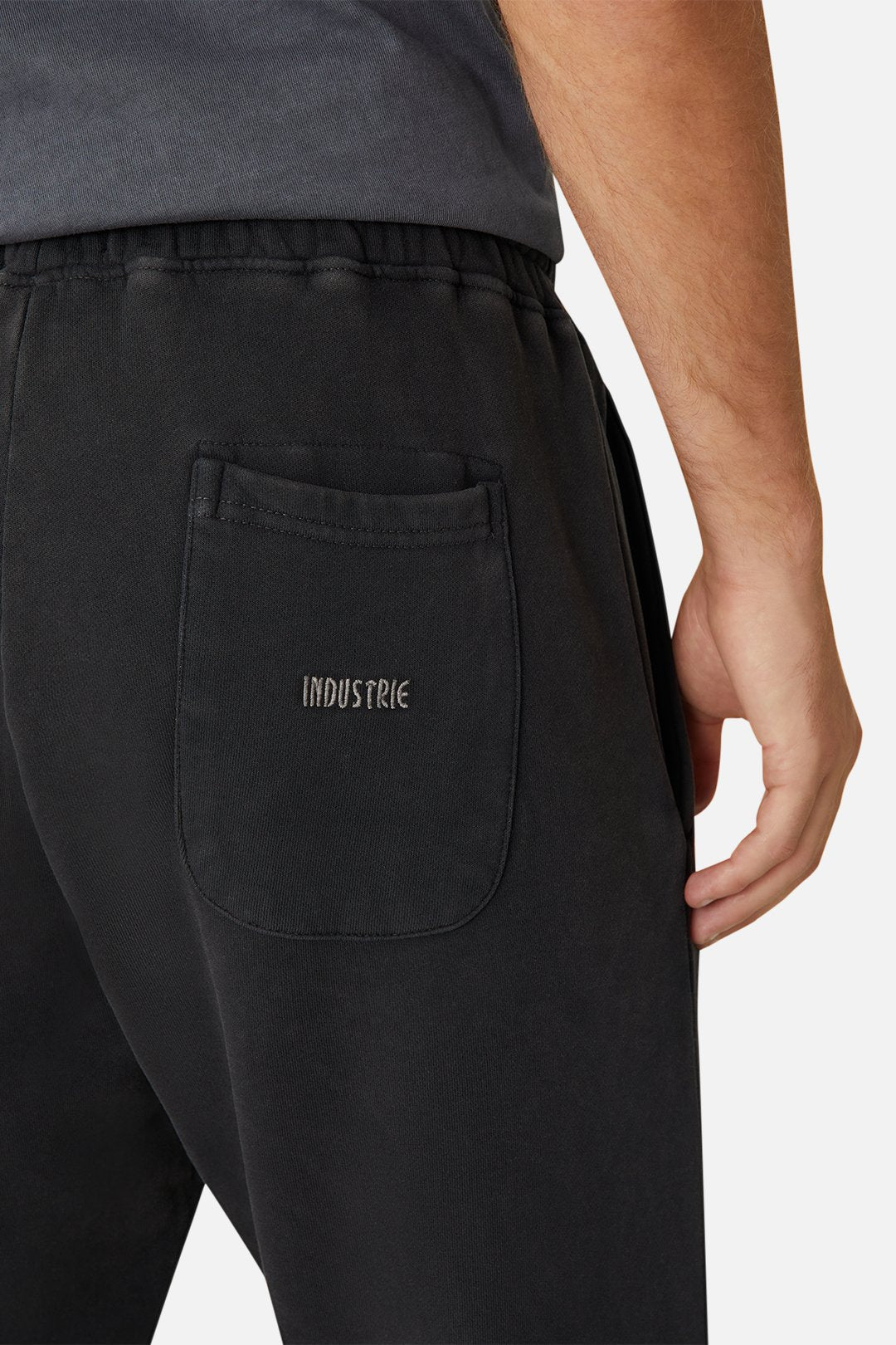 A person wearing the Industrie The Del Sur Track Pant in black, featuring an embroidered logo on the back pocket. Shown from the waist down with one arm partially visible, these track pants have an elastic drawstring waist for enhanced comfort.