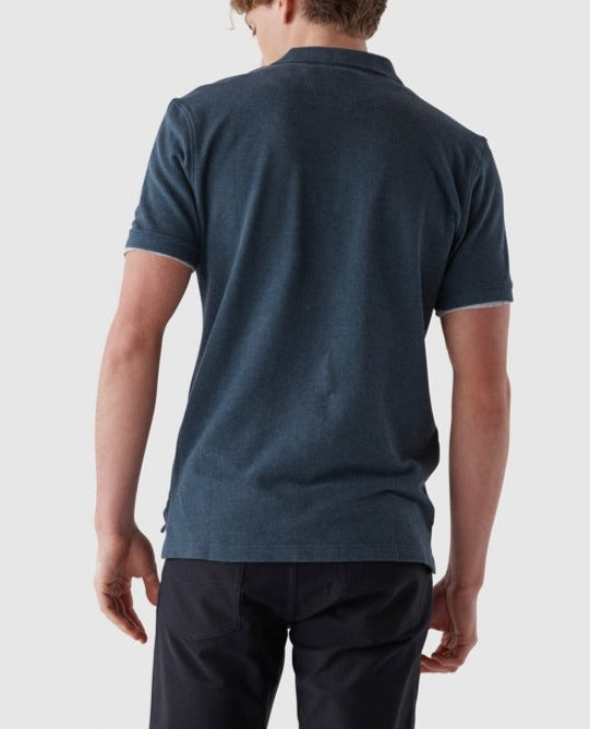 A person with light hair faces away from the camera, wearing a modern fit dark blue Rodd & Gunn The Gunn Polo made of premium Australian Cotton for comfort and style, paired with black pants, against a plain light gray background.