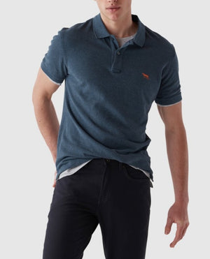 A person in a modern fit Rodd & Gunn The Gunn Polo, blue, made from premium Australian cotton with an embroidered chest logo, paired with black pants against a plain light gray background.