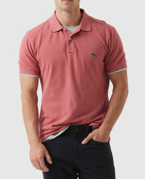 A person wearing a comfortable Rodd & Gunn The Gunn Polo in red, featuring a small animal logo on the left chest pocket, paired with black pants. The modern fit highlights their style as they stand with one hand in their pocket against a plain background. Their face is not visible.