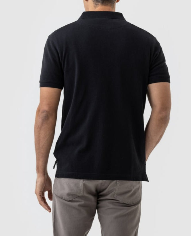 Rear view of a person wearing a black short-sleeved Rodd & Gunn The Gunn Polo, featuring the Rodd & Gunn logo, and light gray pants. They are standing against a plain light gray background.