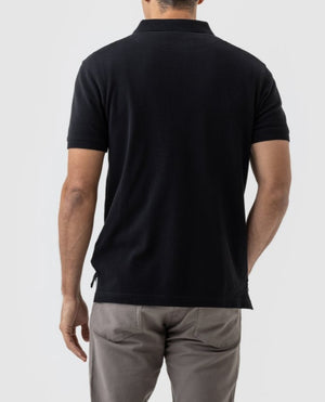 Rear view of a person wearing a black short-sleeved Rodd & Gunn The Gunn Polo, featuring the Rodd & Gunn logo, and light gray pants. They are standing against a plain light gray background.
