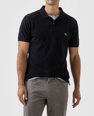 A person is wearing a black Rodd & Gunn The Gunn Polo and gray pants. The polo shirt, made from Australian Cotton, features a small Rodd & Gunn logo embroidered on the left chest. They are standing against a plain, light gray background. The personβs head is not visible in the image.