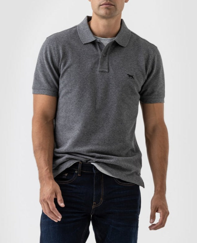 A person wearing a gray Rodd & Gunn The Gunn Polo, featuring a black logo on the chest and crafted from premium Australian cotton, stands against a plain background. The stretch fabric complements their casual pose with dark jeans and one hand slightly raised.