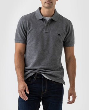A person wearing a gray Rodd & Gunn The Gunn Polo, featuring a black logo on the chest and crafted from premium Australian cotton, stands against a plain background. The stretch fabric complements their casual pose with dark jeans and one hand slightly raised.