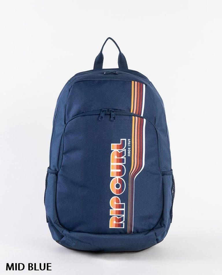 The Rip Curl Ozone 30L Backpack, in dark blue with multiple zippered compartments and adjustable straps, showcases the "RIP CURL" logo prominently in large, colorful vertical text featuring yellow, orange, and red stripes on the front. The background is white and "MID BLUE" is indicated in black text at the bottom left corner.