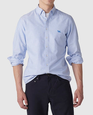 A person wears a light blue Rodd & Gunn Gunn Oxford Sports Fit Shirt made of soft cotton, featuring rolled-up sleeves and a discreet blue logo on the chest, paired with dark pants. The shirt is slightly tucked in at the front against a plain white background.