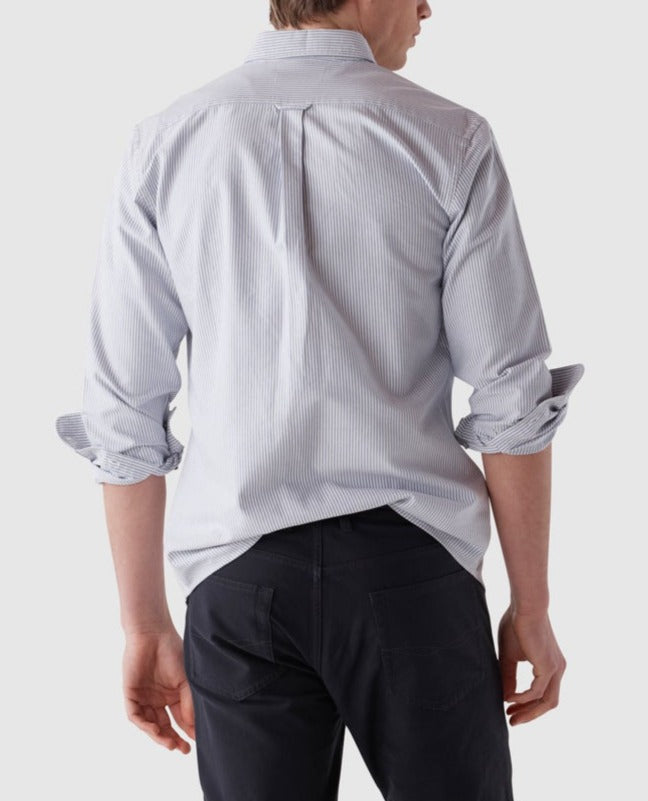 A man stands facing away, wearing a light blue pinstripe Rodd & Gunn Gunn Oxford Stripe Sports Fit Shirt made from 100% cotton, with the sleeves rolled up. He is also wearing dark pants. The background is plain and light grey.