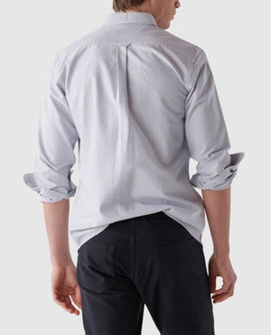 A man stands facing away, wearing a light blue pinstripe Rodd & Gunn Gunn Oxford Stripe Sports Fit Shirt made from 100% cotton, with the sleeves rolled up. He is also wearing dark pants. The background is plain and light grey.