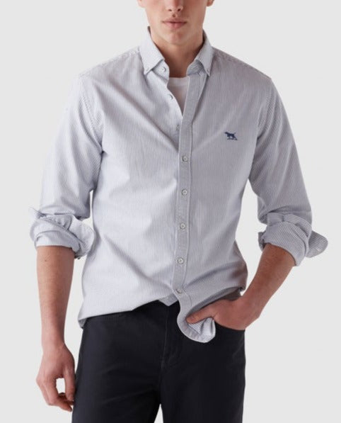 A man stands wearing a light blue Rodd & Gunn Gunn Oxford Stripe Sports Fit Shirt with sleeves rolled up and a small embroidered logo on the left chest. He has one hand casually in his pocket and is paired with dark pants. The sports fit shirt, made from 100% cotton, complements the plain, light-colored background perfectly.