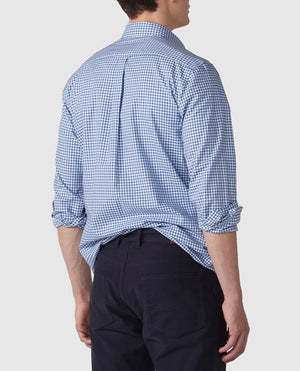 A man is photographed from behind, wearing the stylish Rodd & Gunn Gunn Check Oxford Sports Fit Shirt, which has a blue and white-checkered pattern. The shirt, made of 100% cotton, has its sleeves rolled up and is loosely tucked in at the back. He pairs it with dark-colored pants against a plain, light grey background.