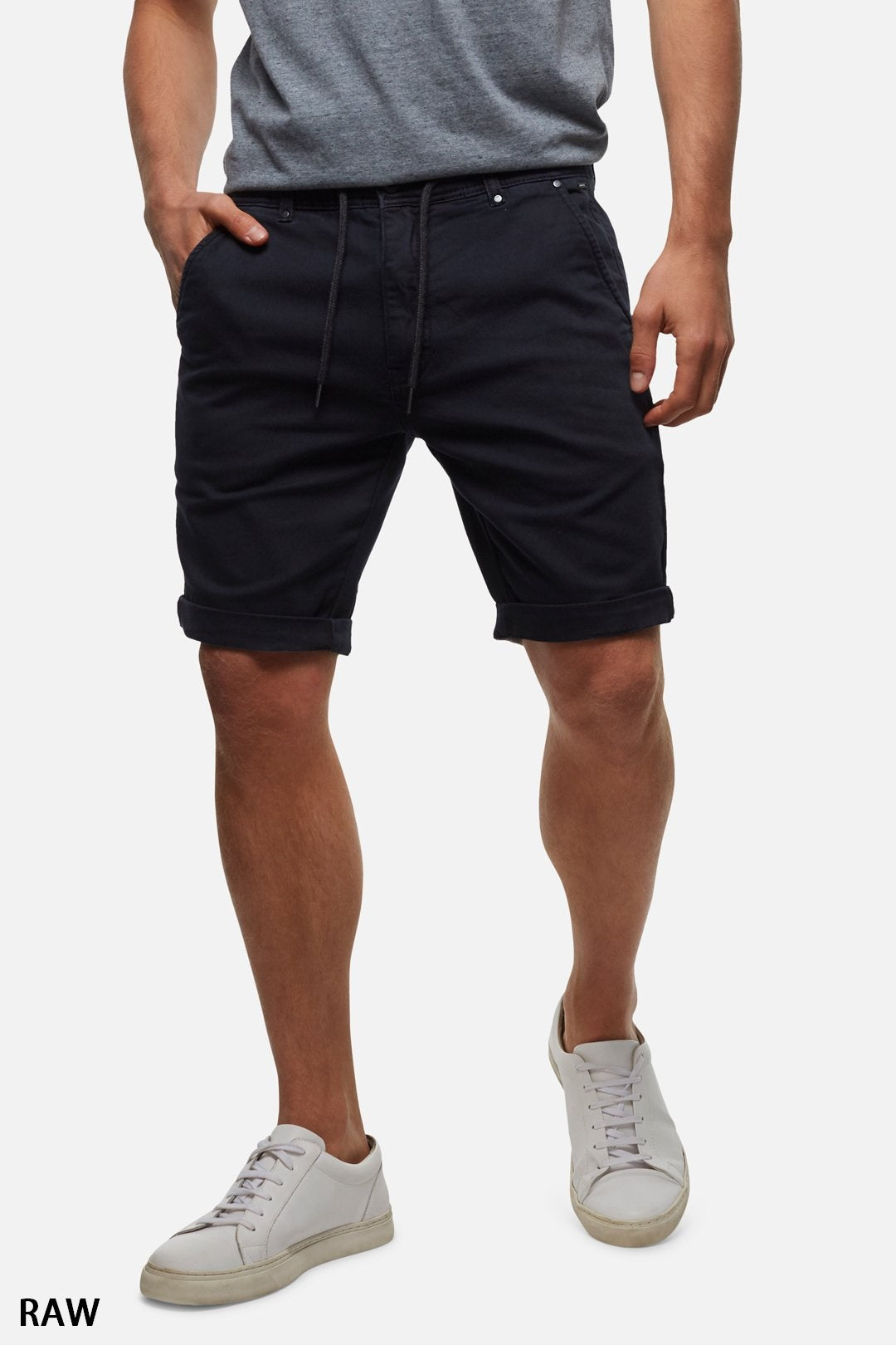 A person is shown from the shoulders down, wearing a gray t-shirt, Industrie The Drifter Cuba Short in modern slim fit navy blue with a drawstring, and white sneakers. The person is standing with their hands in their pockets against a plain white background. The text "RAW" appears in the bottom left corner.