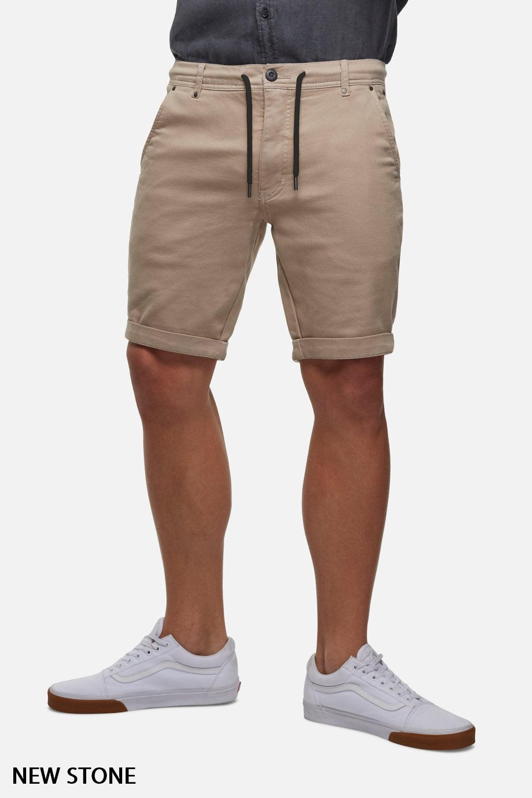 Person wearing Industrie The Drifter Cuba Short in a light brown hue, with a comfortable fit and a black drawstring. They have paired it with a tucked-in grey button-up shirt and white sneakers featuring brown soles. Text at the bottom left corner reads "NEW STONE." The background is white.