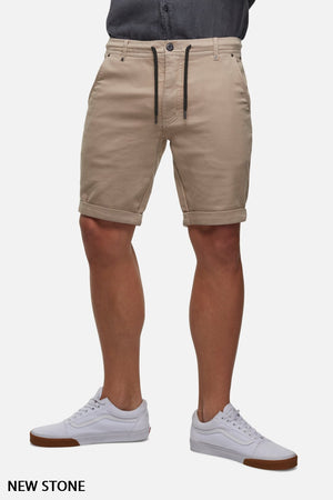 Person wearing Industrie The Drifter Cuba Short in a light brown hue, with a comfortable fit and a black drawstring. They have paired it with a tucked-in grey button-up shirt and white sneakers featuring brown soles. Text at the bottom left corner reads "NEW STONE." The background is white.
