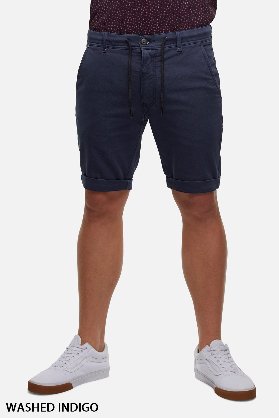A person is wearing the Industrie The Drifter Cuba Shorts in a washed indigo, featuring a modern slim fit and knee-length, with a drawstring waist. The shorts are paired with a dark top and white sneakers. The background is plain white. Text at the bottom left corner reads "WASHED INDIGO.