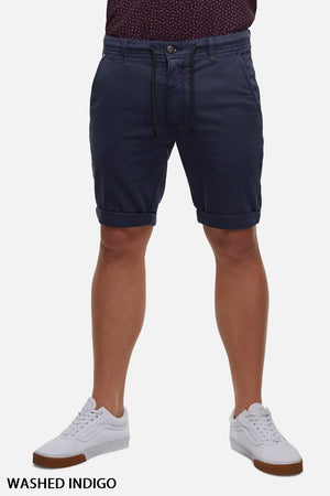 A person is wearing the Industrie The Drifter Cuba Shorts in a washed indigo, featuring a modern slim fit and knee-length, with a drawstring waist. The shorts are paired with a dark top and white sneakers. The background is plain white. Text at the bottom left corner reads "WASHED INDIGO.