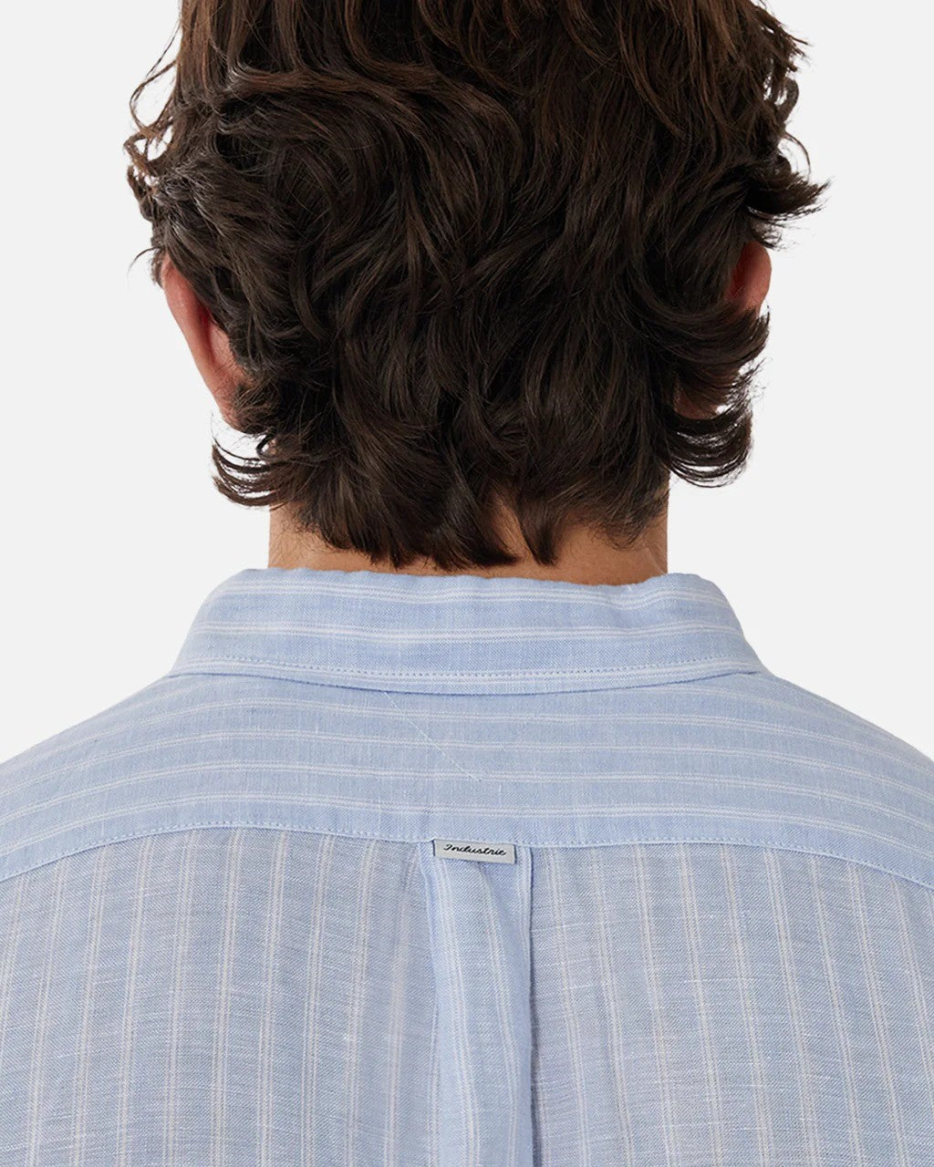 The image showcases the back of a person with wavy, medium-length hair, wearing the Industrie The Mattera Long Sleeve Shirt in light blue with subtle vertical stripes. A small tag adorns the upper back of this stylish and comfortable linen shirt.