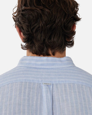 The image showcases the back of a person with wavy, medium-length hair, wearing the Industrie The Mattera Long Sleeve Shirt in light blue with subtle vertical stripes. A small tag adorns the upper back of this stylish and comfortable linen shirt.