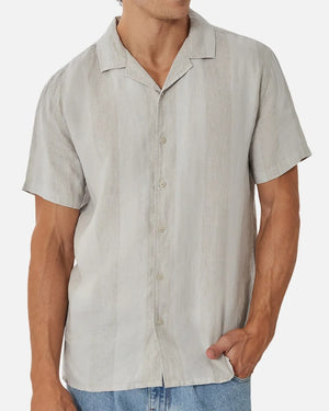 A person wears the Industrie The Kanaki Short Sleeve Shirt in light gray, featuring subtle vertical stripes, paired with light blue jeans and stands with one hand in their pocket against a plain background.