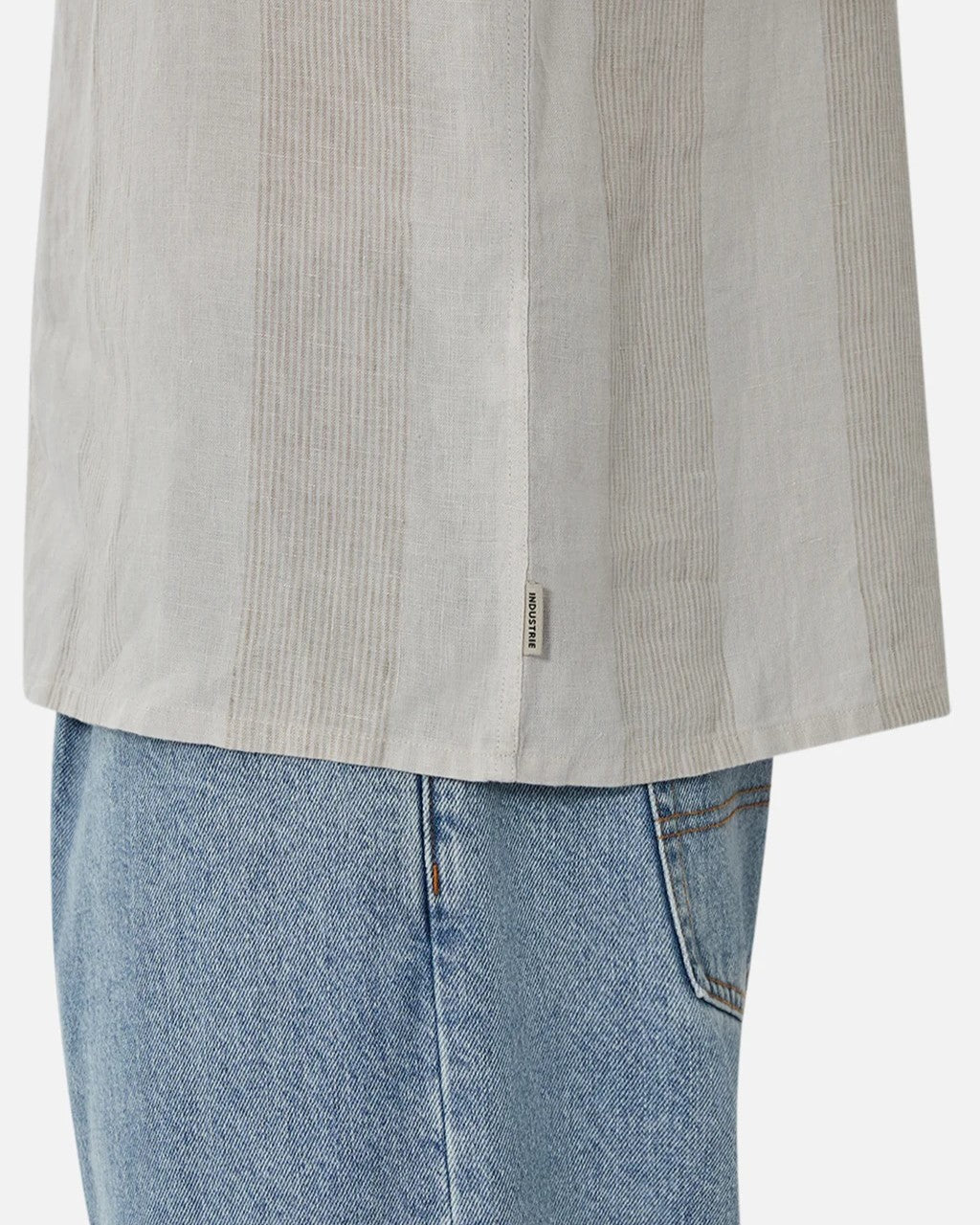 Close-up of a person wearing the Industrie The Kanaki Short Sleeve Shirt with light vertical stripes, displaying a tag on the side, paired with light blue jeans. The focus is on the lower half of the shirt and top of jeans.