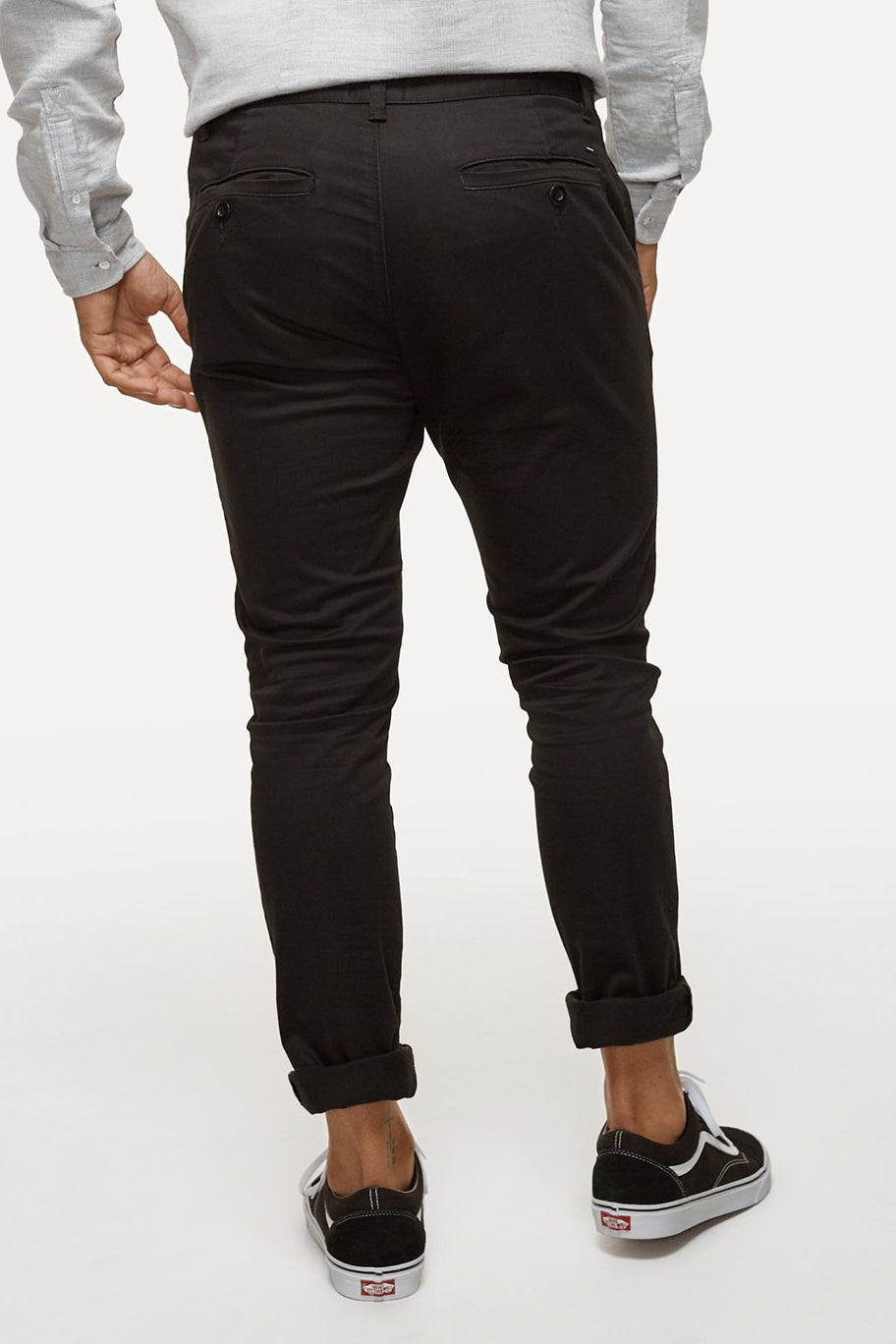 A person is seen from the back wearing black Industrie The Cuba Chino Pant, a light gray long-sleeve shirt, and black Vans slip-on shoes with white soles. The pants are slightly rolled up at the ankles. The background is off-white.