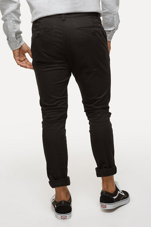 A person is seen from the back wearing black Industrie The Cuba Chino Pant, a light gray long-sleeve shirt, and black Vans slip-on shoes with white soles. The pants are slightly rolled up at the ankles. The background is off-white.