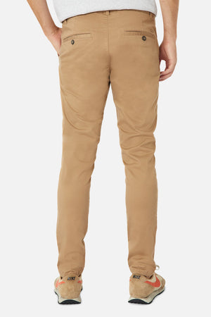 A person is wearing Industrie The Cuba Chino Pant in a skinny fit, seen from the back with hands in pockets. They're paired with a light gray shirt and beige Nike sneakers with orange and red details. Against the plain white background, this cotton outfit makes a statement.