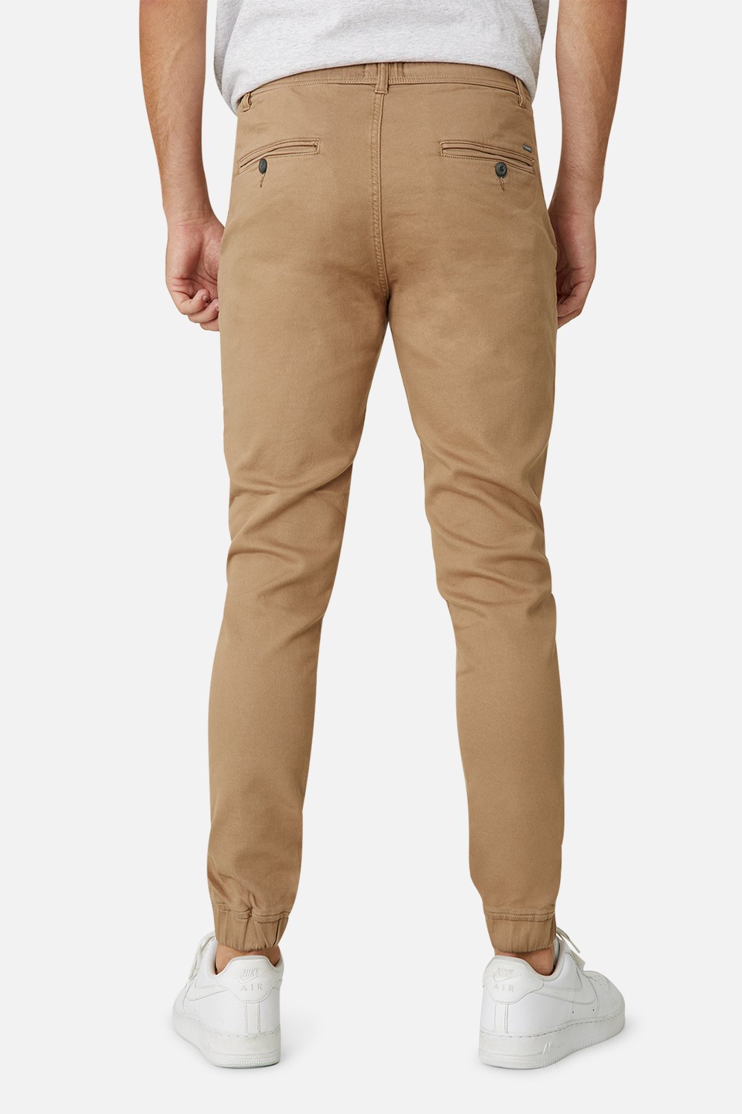 A person is seen from behind in Industrie The Drifter Chino Pant, tan jogger style with an elastic waist and cuffs, paired with a slim-fit white T-shirt. The joggers feature buttoned back pockets and are complimented by sleek white sneakers.