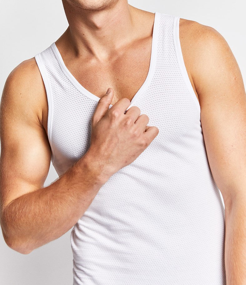 A person wearing a Bonds Coral Island Aths Singlet in white is shown from the shoulders down. The person's left hand is touching their upper chest while their arm is slightly flexed, demonstrating the improved shape retention of the fabric. The background is plain white.