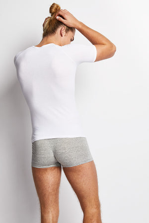 A person with long hair tied in a bun is shown from the back, facing a white wall. They are wearing a Bonds New Coral Island V-Neck Tee in white and light gray boxer briefs. Their left hand touches the back of their head while their right arm hangs loosely at their side, showcasing the breathability of their outfit.