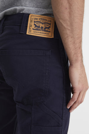 A close-up view of a person wearing dark blue Levis Workwear 505 Utility Pants with durable oversized pockets. The back pocket, Levi's signature stitching, and branded brown leather patch on the waistband are visible. The person is wearing a gray T-shirt, and their hand is positioned by their side.