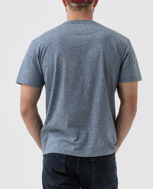 A person wearing a Rodd & Gunn The Gunn T-Shirt in heather blue, paired with dark blue jeans and a red cap, stands with their back facing the camera against a light grey plain background.