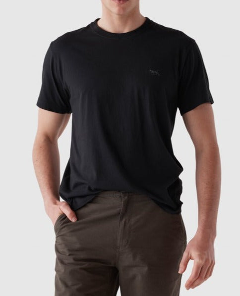 A person wearing a plain black Rodd & Gunn The Gunn T-Shirt made from Australian Cotton and brown pants stands against a neutral gray background. Their right hand is in their pants pocket, and the left hand hangs by their side. Their face is not visible in the image.