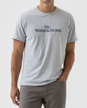 A person is wearing a Rodd & Gunn Logo T-Shirt, a light gray sports fit tee made from Australian Cotton, featuring the brand's logo on the chest. They have one hand in the pocket of their matching gray pants and are standing against a plain white background.