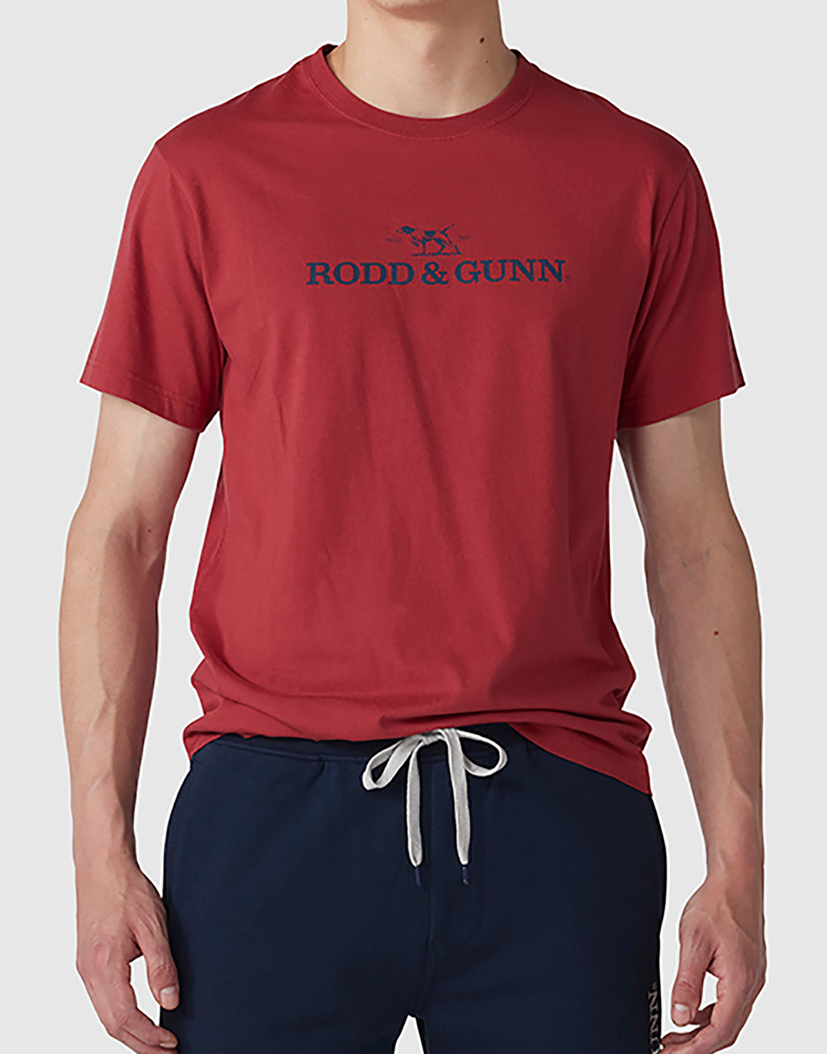 A person is wearing a red Rodd & Gunn Logo T-Shirt featuring the text "RODD & GUNN" and a logo with a dog. They are also sporting navy blue pants with a white drawstring. The background is plain light gray.