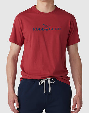A person is wearing a red Rodd & Gunn Logo T-Shirt featuring the text "RODD & GUNN" and a logo with a dog. They are also sporting navy blue pants with a white drawstring. The background is plain light gray.
