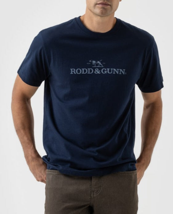 Wearing a navy Rodd & Gunn Logo T-Shirt made from soft Australian cotton, the man showcases its iconic small dog logo. With his hands in brown pants' pockets, he exudes a confident, refined look against a plain backdrop.