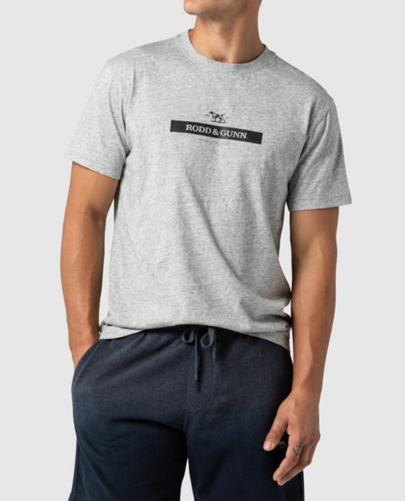 A person wearing a Rodd & Gunn Cumulus Sports Fit Tee in light grey, made of premium cotton and featuring the Rodd & Gunn logo in black across the chest, paired with dark navy drawstring pants. The individual is posing with one hand in their pocket against a plain light grey background.