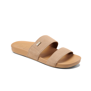 The REEF Cushion Vista Suede is a tan slide sandal with vegan leather straps, a small metallic tag on the upper strap, and a contoured footbed. It features a cushion bounce midsole and ridged sole for grip.