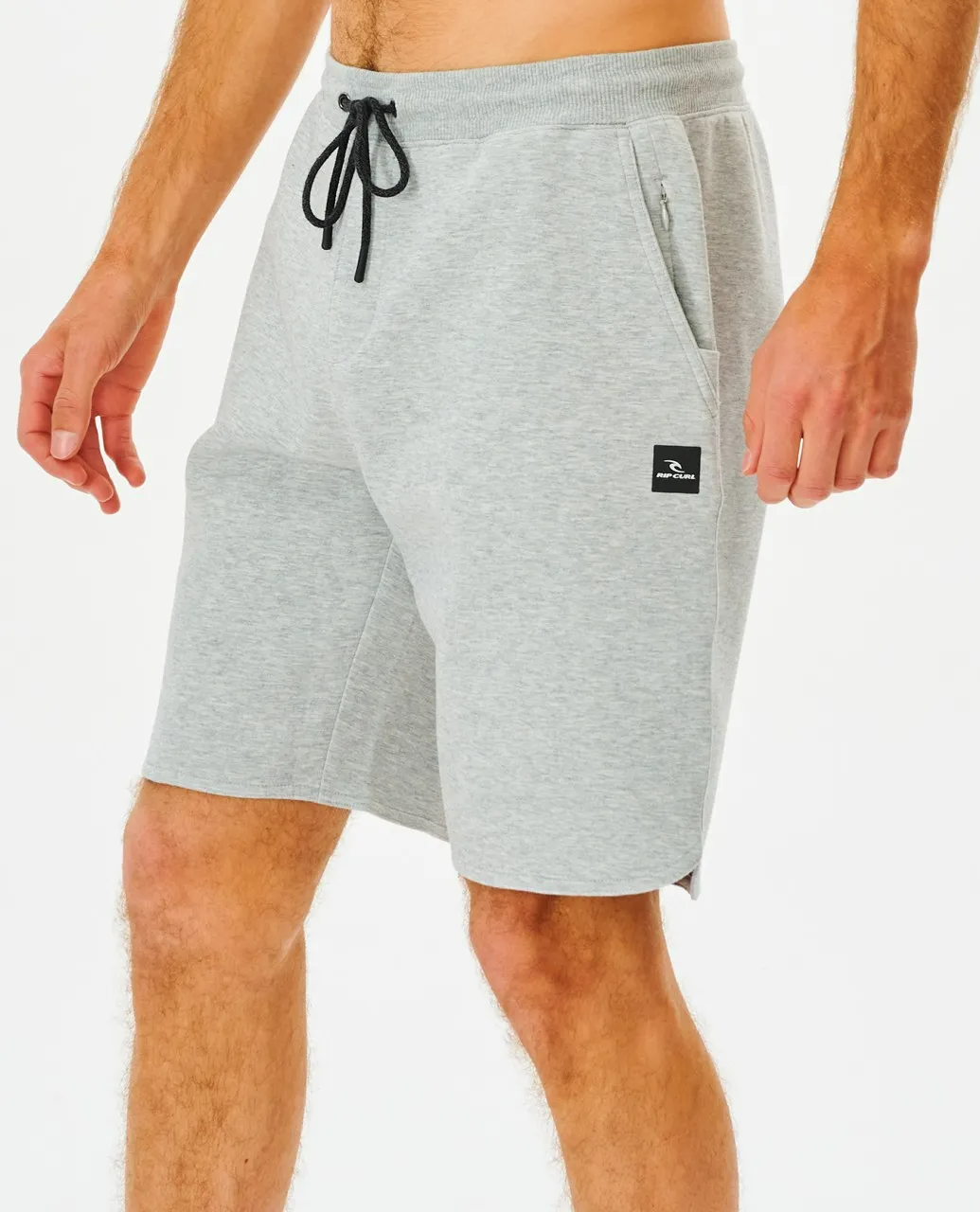 A person wears Rip Curl Vaporcool Shorts in light gray with a black drawstring and logo on the left leg. These comfy shorts have side pockets, perfect for lounging or workouts, shown against a white backdrop.