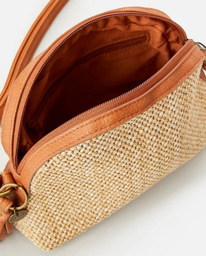 The Rip Curl Surf Gypsy Cross Body Bag boasts a tan straw texture with rust leather accents, a zipper closure, adjustable shoulder strap, and detachable tassel keyring that adds flair to its partially open view, revealing the fabric-lined interior.