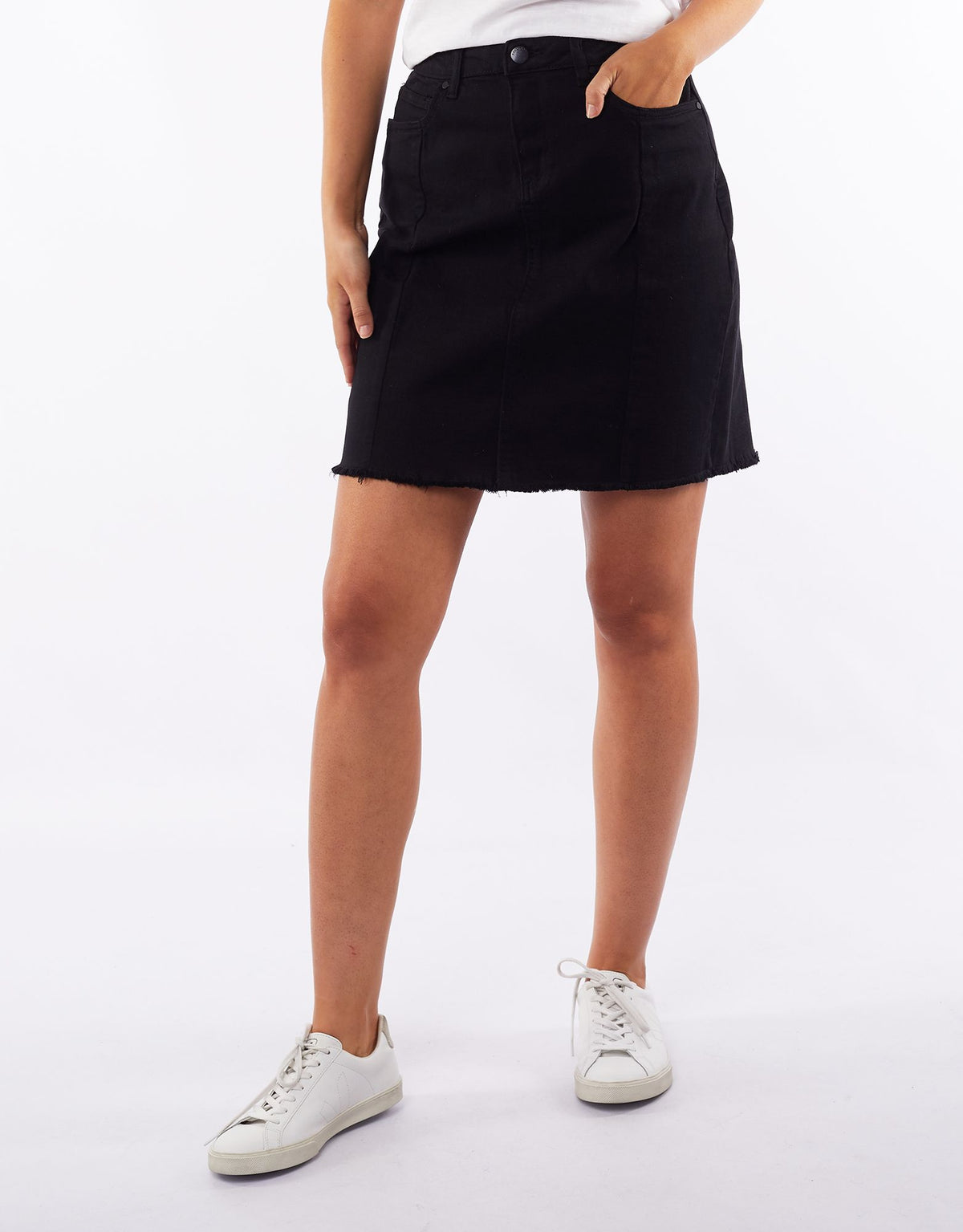 A person is standing against a white background, wearing the Foxwood Kiama Skirt, which features black denim with frayed detailing at the hem. They are also wearing a white top and white sneakers, with their right hand placed in the pocket of the skirt.